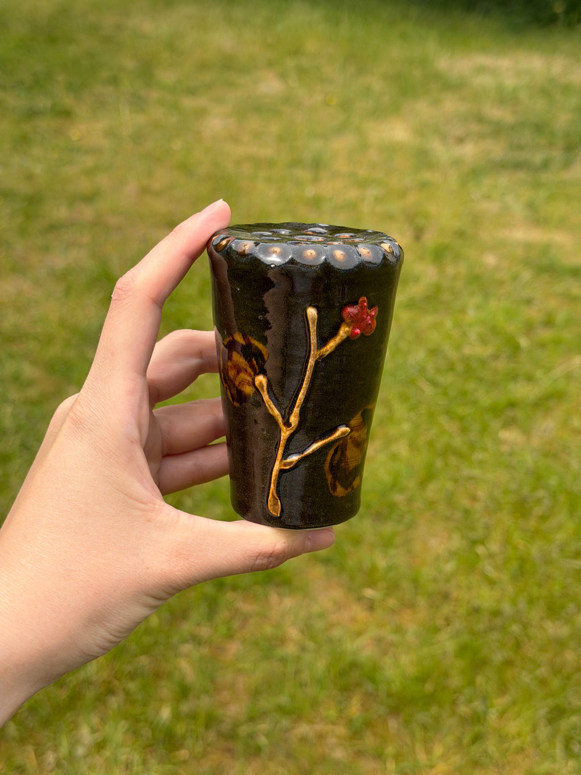 Hand holding a black ceramic vase with floral designs against a grassy background