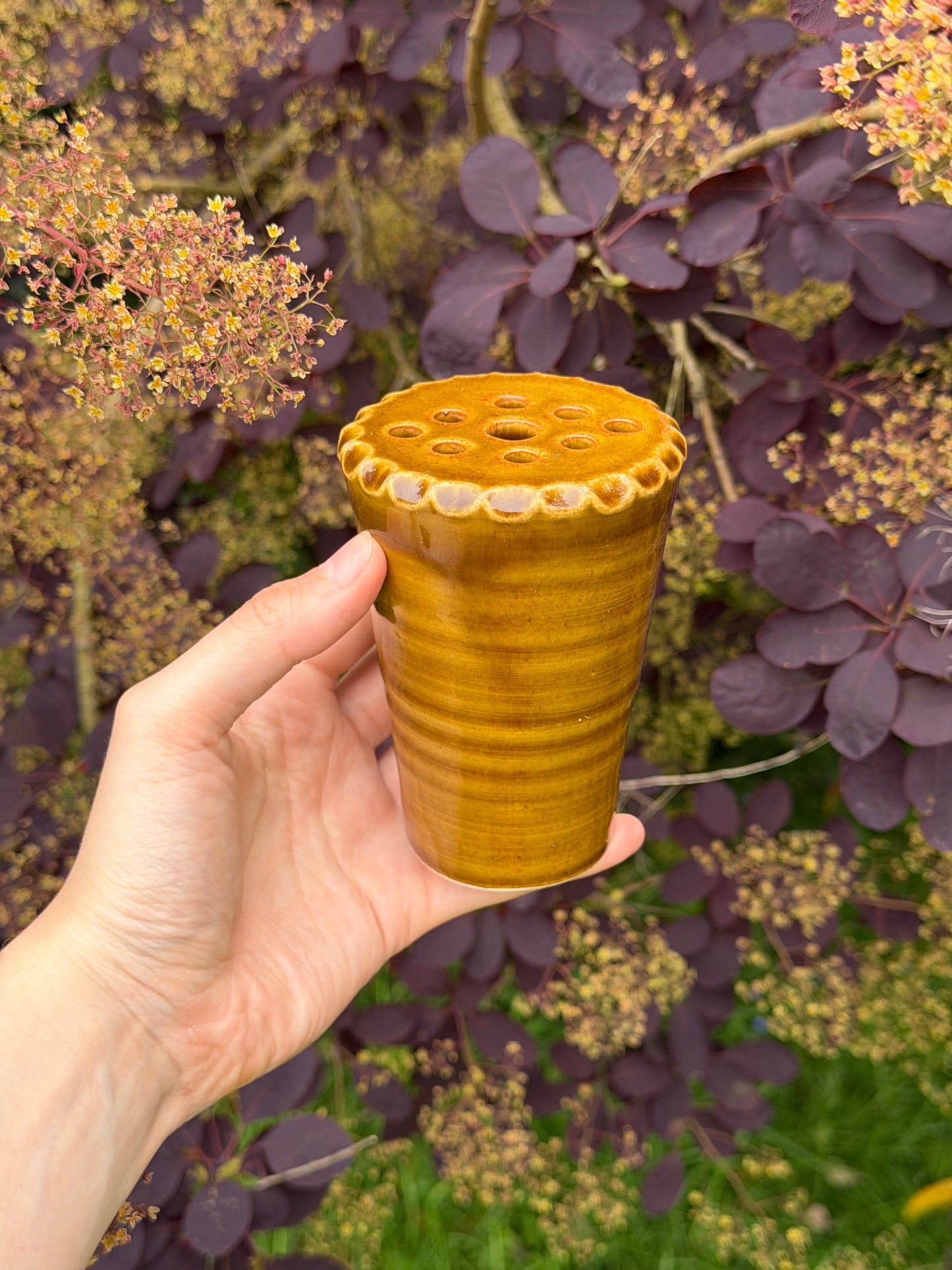 Hand holding a textured yellow cup against a natural background with purple foliage.